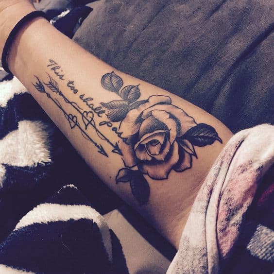 A "This Too Shall Pass" tattoo in cursive, with a rose and arrow designs, on a person's forearm.