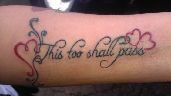 A "This Too Shall Pass" tattoo on a person's forearm, with cursive lettering and decorative red heart and flower details.