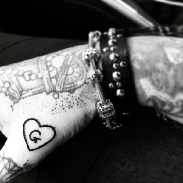 Small tattoo of a heart with the letter “G” inside on the thumb, symbolizing love and devotion to someone special.