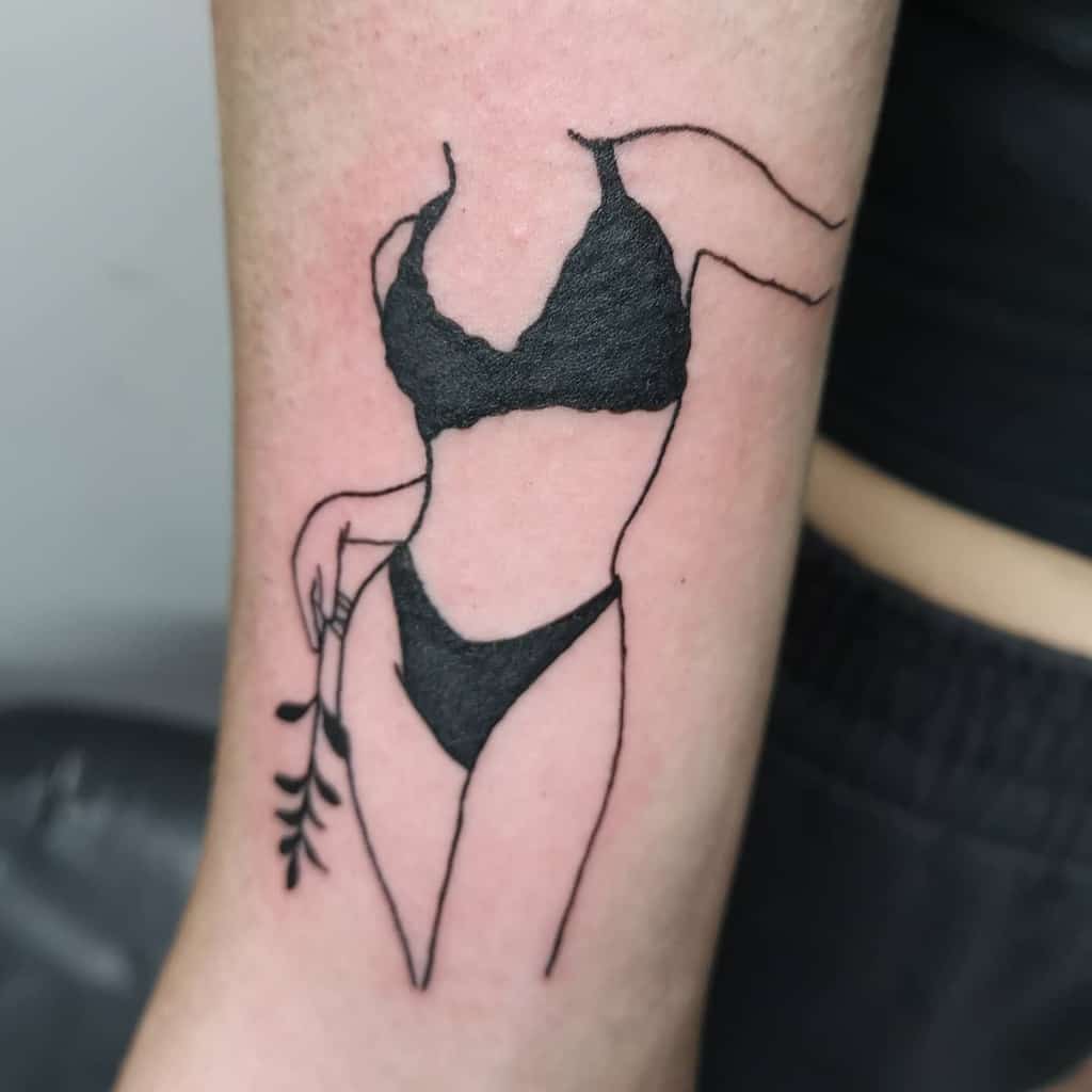 Minimalist black ink tattoo of a woman in a bikini, outlined with fine lines, holding a small branch, with bold shading on the swimsuit