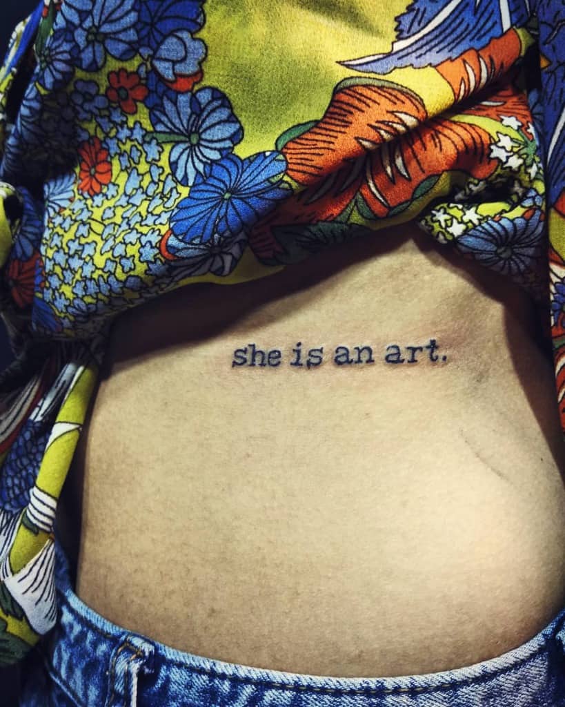 Simple black ink text tattoo on the ribcage reading 'she is an art.' in a typewriter-style font
