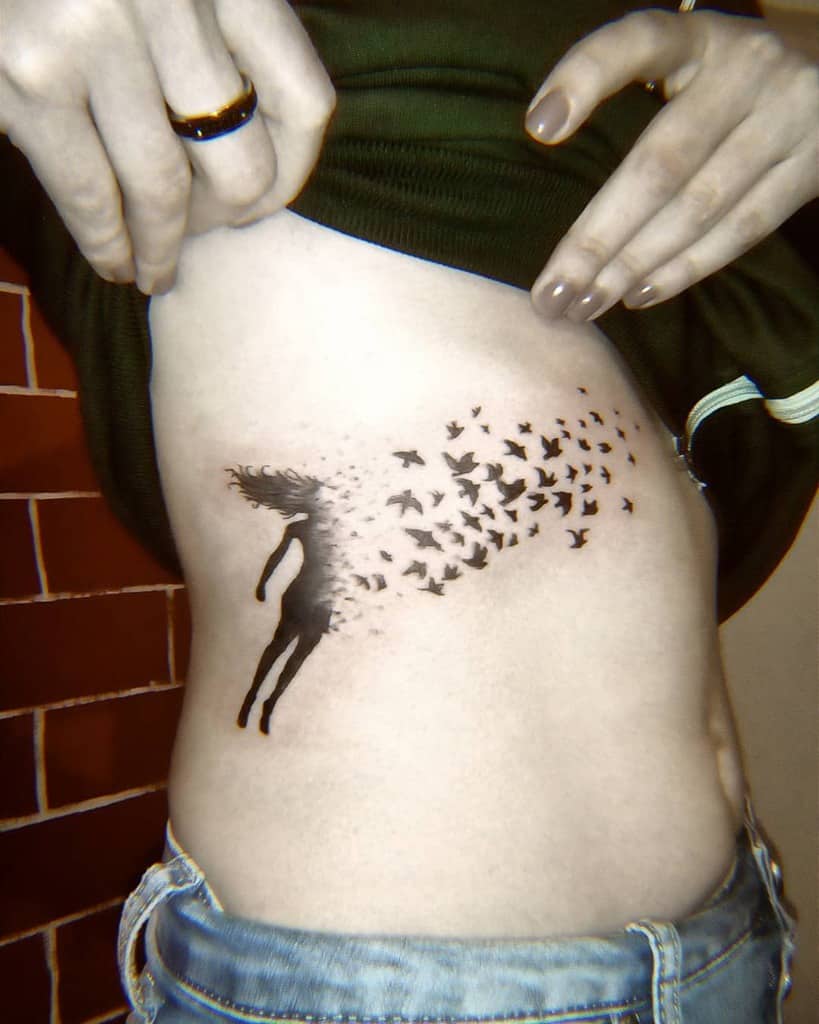 Black ink tattoo of a silhouette of a woman dissolving into a flock of birds in flight, symbolizing freedom and transformation