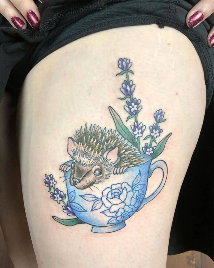A colorful tattoo on the thigh featuring a hedgehog peeking out of a blue floral teacup with lavender sprigs on top.