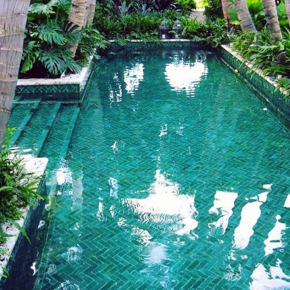 Tropical pool with emerald-green herringbone tiles surrounded by lush greenery and palm trees.