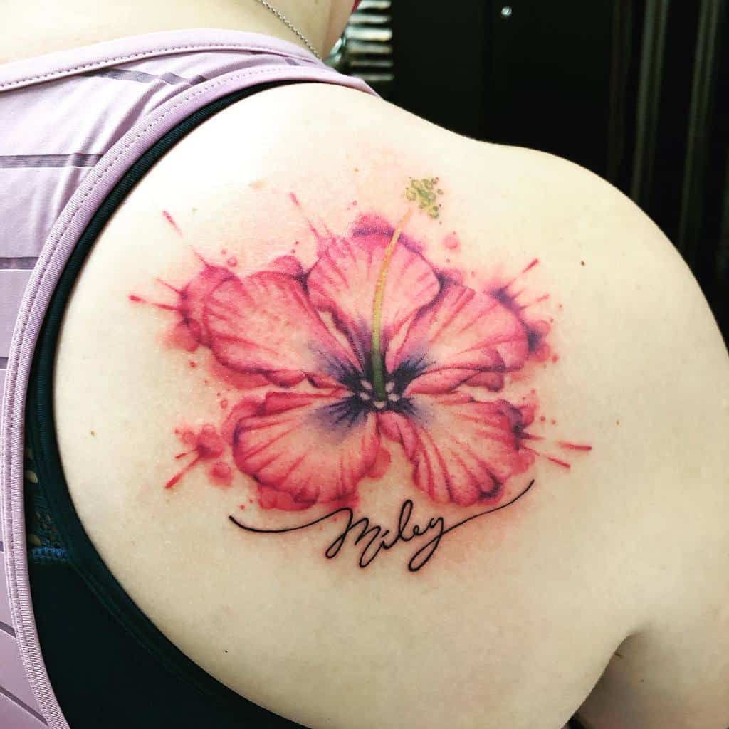 A watercolor hibiscus tattoo graces the shoulder, with splashes of pink and purple hues and elegant cursive text flowing below