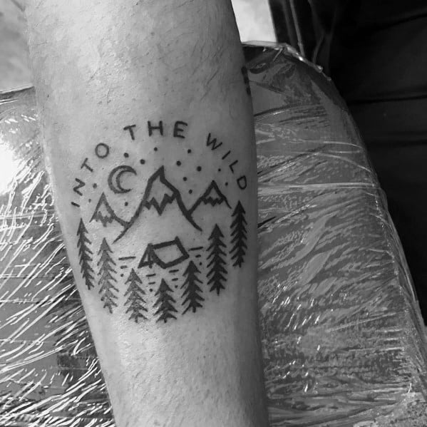 A black and gray minimalist tattoo of mountains, trees, a tent, and the phrase "Into the Wild," symbolizing adventure and a love for nature.