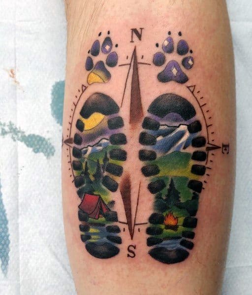 This tattoo features colorful boot prints filled with nature scenes, a compass, paw prints, and camping elements, symbolizing adventure and exploration.