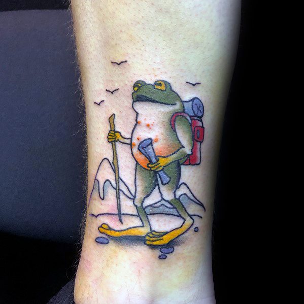 Colorful tattoo of a hiking frog with a backpack, trekking pole, and flashlight, set against a mountain backdrop, symbolizing adventure.