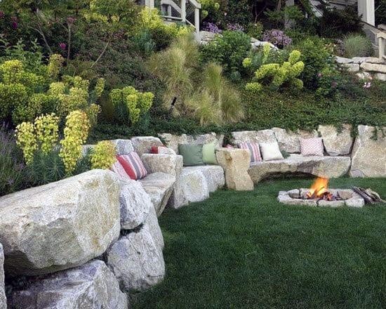 Sloped backyard with a cozy fire pit area, built-in stone seating, lush greenery, and a giant rock retaining wall for natural charm