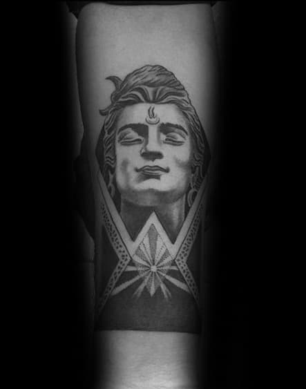 Forearm tattoo of Lord Shiva with a serene, upward gaze, geometric elements framing the face, and a radiant starburst below.