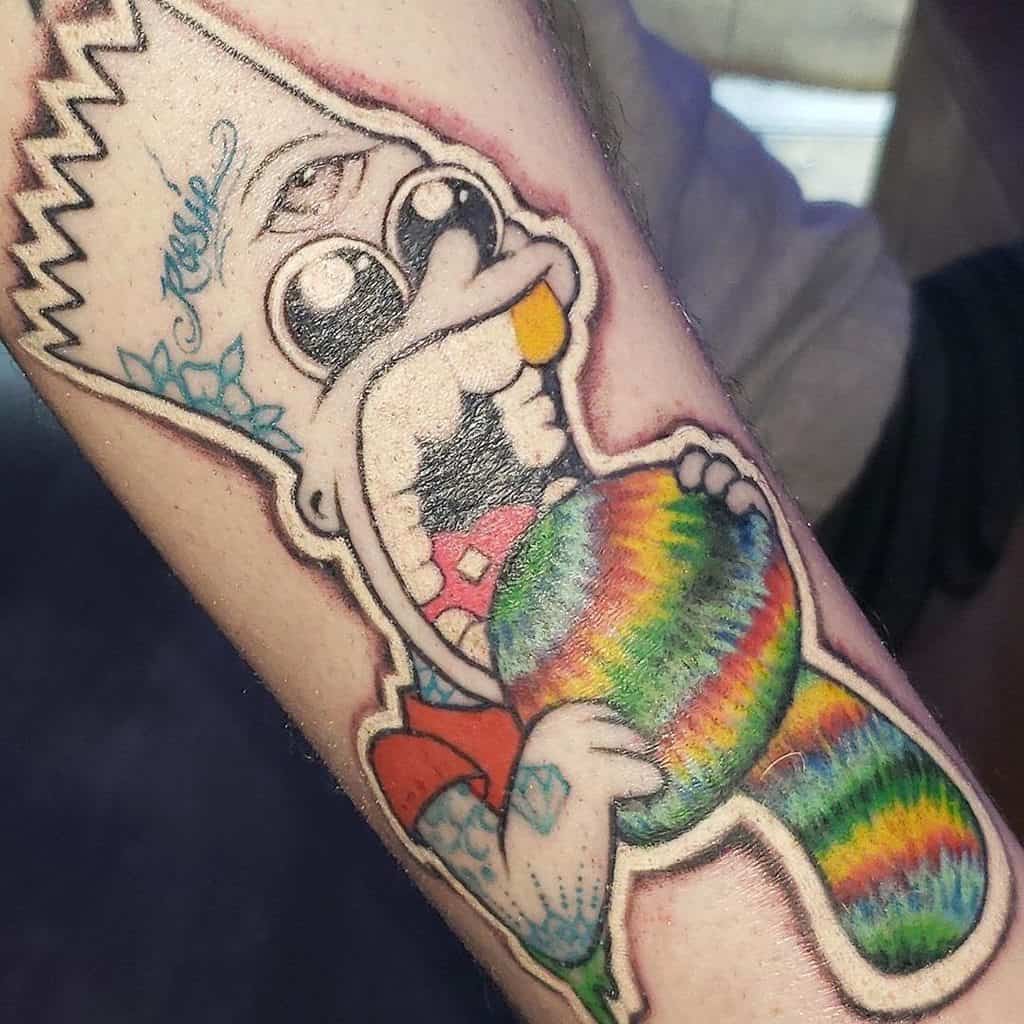 Colorful tattoo of a cartoon character with a third eye, exaggerated features, and a rainbow lollipop, inked on the forearm.