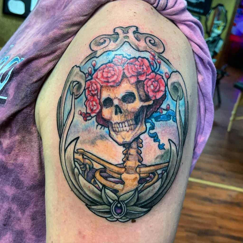 Ornate tattoo of a skeleton with a rose crown inside a decorative frame, blending gothic and vintage art styles on the upper arm.