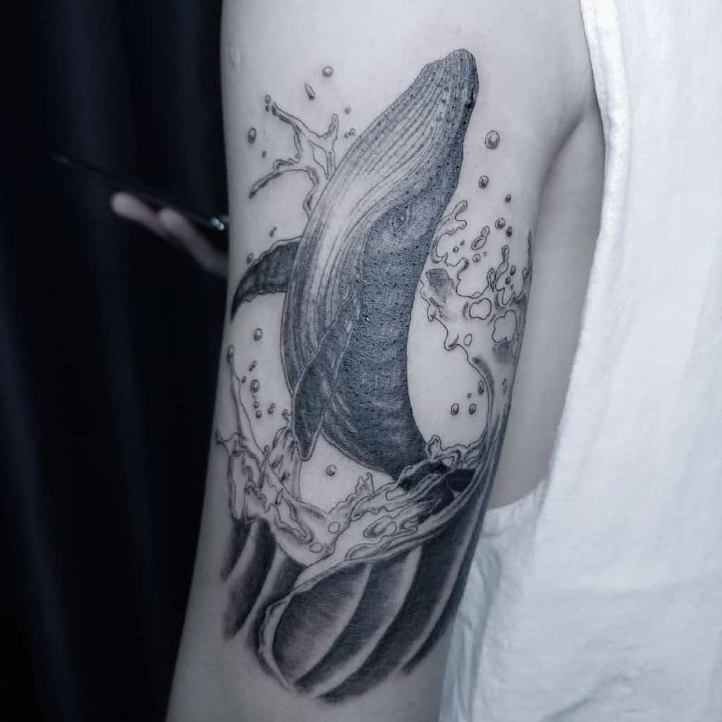 Black and white tattoo of a whale jumping out of water on an upper arm
