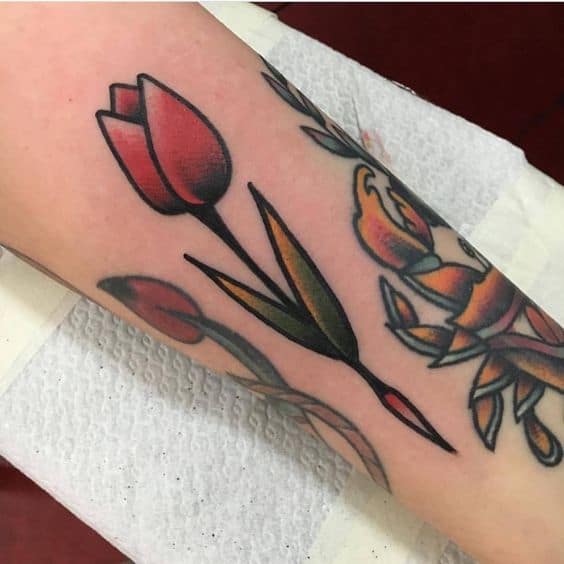 A bold neo-traditional tulip tattoo on the forearm, featuring a red flower with dark shading, green leaves, and crisp black outlines.