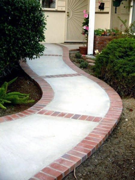 A curved walkway with red brick edges leads to a doorway, surrounded by lush greenery
