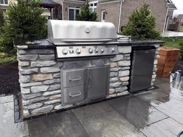 Outdoor stone grill with built-in grill, stainless doors, and fridge on a wet patio