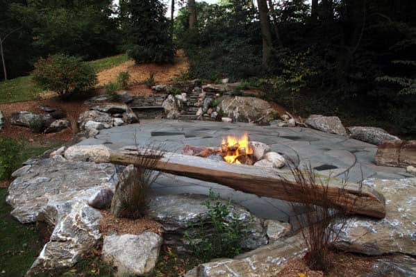 A stone patio with a burning fire pit, surrounded by trees and rocks in a forest setting