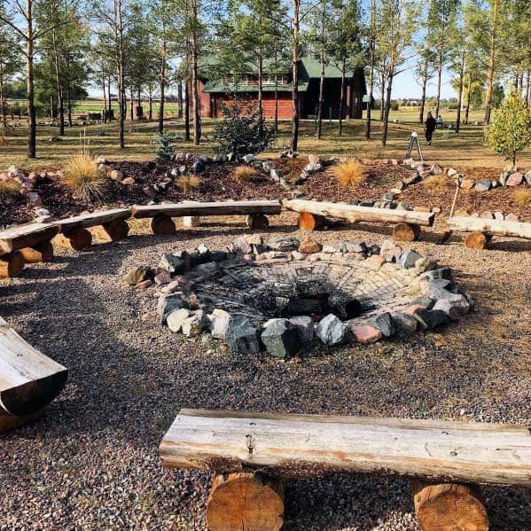 Wooden benches encircle a stone firepit in a park, with a rustic cabin and trees in the background