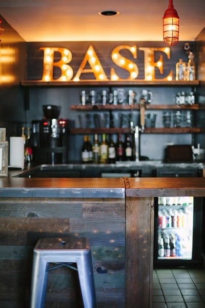 Cozy bar with a wooden counter, metal stools, and an illuminated "BASE" sign above shelves filled with glassware and bottles