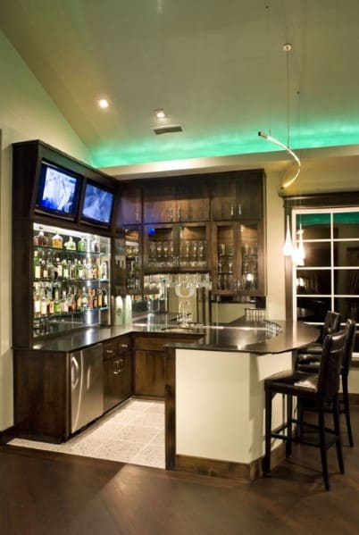 Modern home bar with dark wood cabinets, illuminated shelves, two TVs, and a sleek black countertop with high chairs