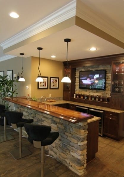 Stone-counter home bar with wooden top, pendant lights, dark chairs, and a wall-mounted TV