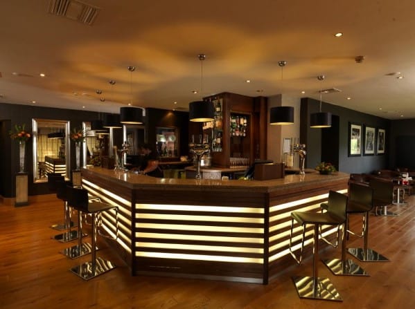 A modern bar with an illuminated counter, swivel chairs, and ambient lighting in a warmly decorated space