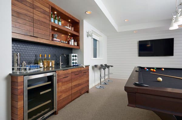Modern game room with a pool table, wall-mounted TV, and wooden bar featuring a wine cooler and shelves