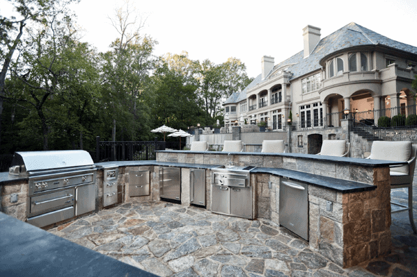 Outdoor kitchen with a built-in grill, stainless steel appliances, and views of a large mansion surrounded by trees