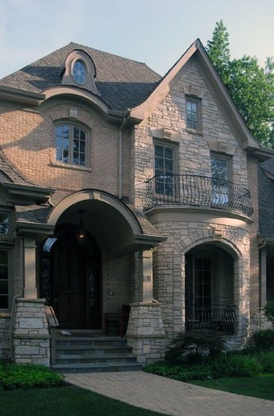 Elegant home with a mix of light brick and stone cladding, arched entryway, wrought iron balcony, and intricate architectural details