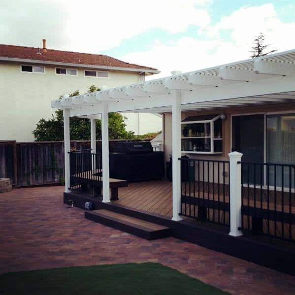 Backyard patio with white pergola, black railing, and brick flooring, bench by the grill near large windows