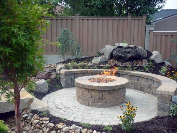 A backyard with a circular stone fire pit, surrounded by plants and wooden fencing
