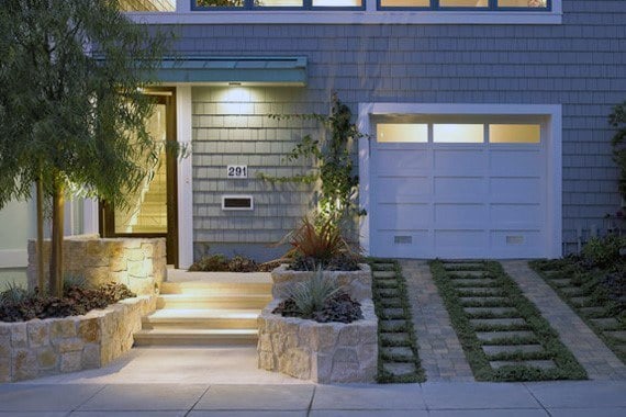 Illuminated modern home entrance with stone steps, greenery, and a white garage with grass pavers.