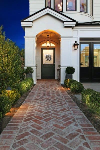 Charming brick walkway with a herringbone pattern, flanked by neatly trimmed hedges leading to a well-lit front door