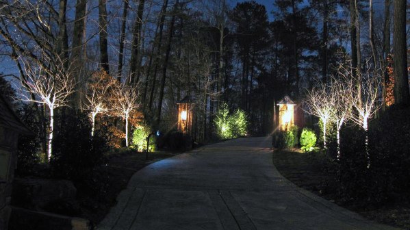 Driveway lined with tree uplighting and lantern-style fixtures, casting a soft glow and creating an inviting atmosphere at night