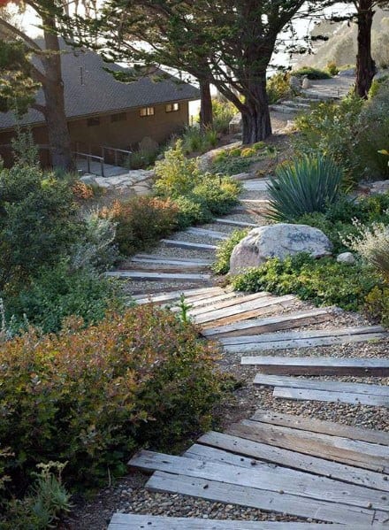 Rustic sloped landscape featuring a wooden plank pathway, gravel steps, lush greenery, and mature trees for a natural woodland look
