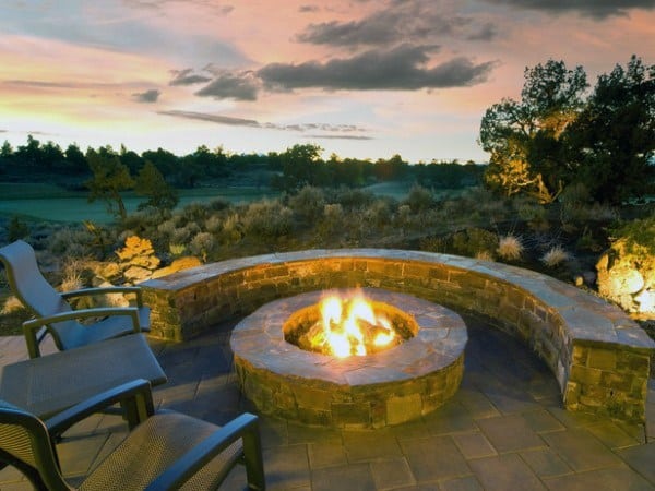 Fire pit with stone seating and chairs, set against a sunset landscape with trees and shrubs in the background