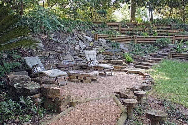 Outdoor stone patio with lounge chairs, surrounded by trees and terraced garden beds on a sloped hill