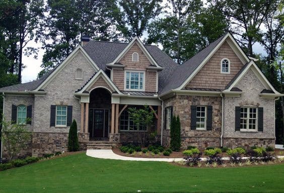 Elegant home with a mix of brick, stone, and shingle cladding, gabled rooflines, a welcoming porch, and lush landscaped front yard