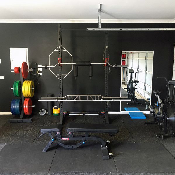 Home gym with black walls, rubber flooring, bench press, barbell rack, and colorful weights.