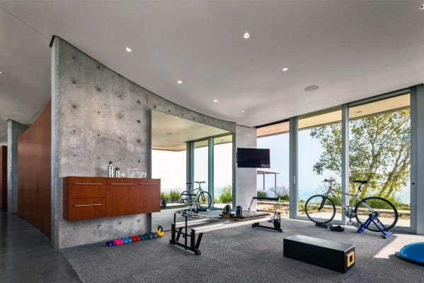 Modern home gym with large windows, exercise bikes, rubber flooring, and a concrete accent wall.