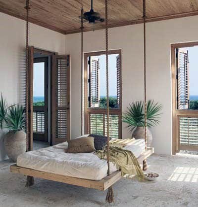 Tropical hanging bed with rope supports, light bedding, and large windows with wooden shutters.