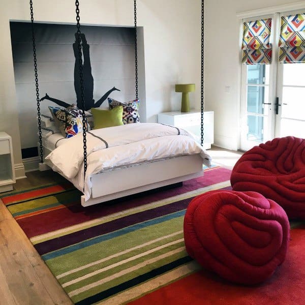 Modern hanging bed with chains, colorful decor, bold rug, and unique red chairs in a bright room.