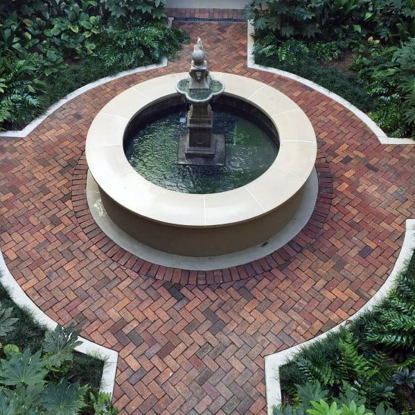 A circular fountain graces a brick courtyard with a walkway surrounded by greenery