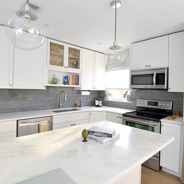 Contemporary kitchen with white cabinets, a grey stone backsplash, marble island, and glass pendant lighting