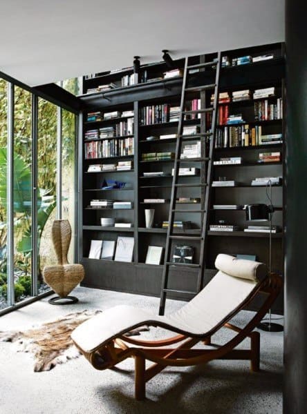 Modern room with bookshelves, a lounge chair, ladder, and large glass windows offering a view of greenery outside