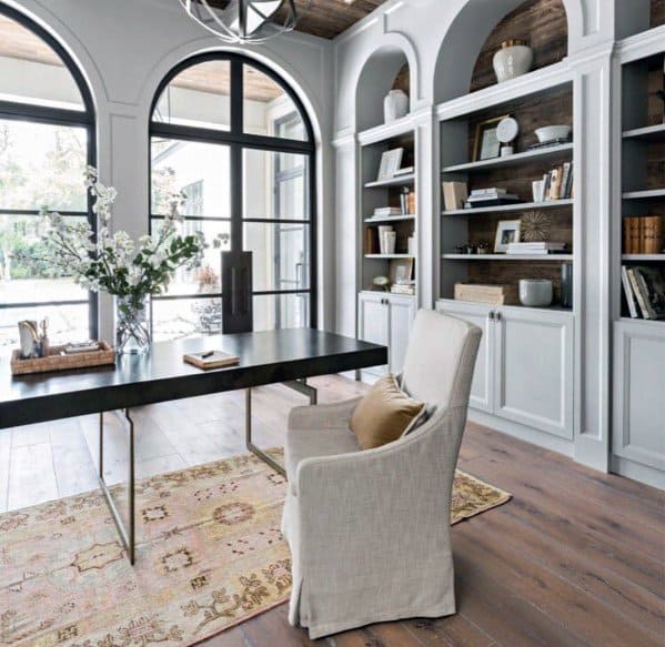 Elegant home office with arched built-in bookcases, black desk, large windows, and a vintage rug on wood flooring