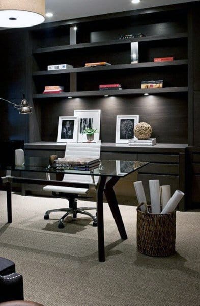Contemporary home office with glass desk, dark built-in shelves, and soft lighting.