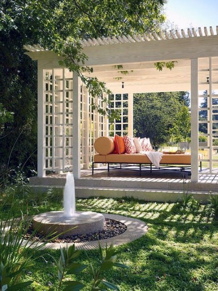 White pergola with lattice walls, vibrant seating, and a small fountain in a lush garden setting.