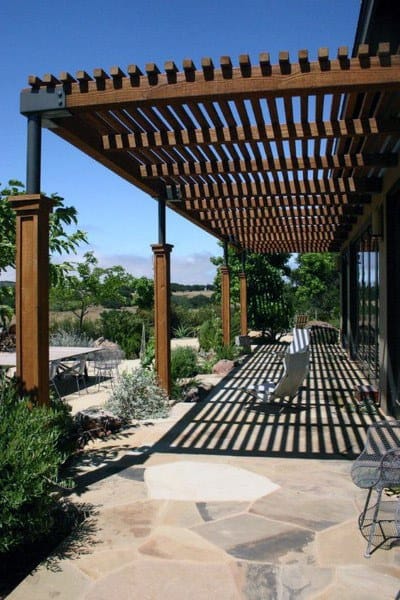 Wooden pergola with slatted roof, stone patio, and outdoor seating overlooking a garden.