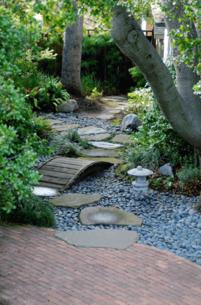 Serene garden path with river rocks, stepping stones, and a wooden bridge, surrounded by lush greenery and a decorative lantern
